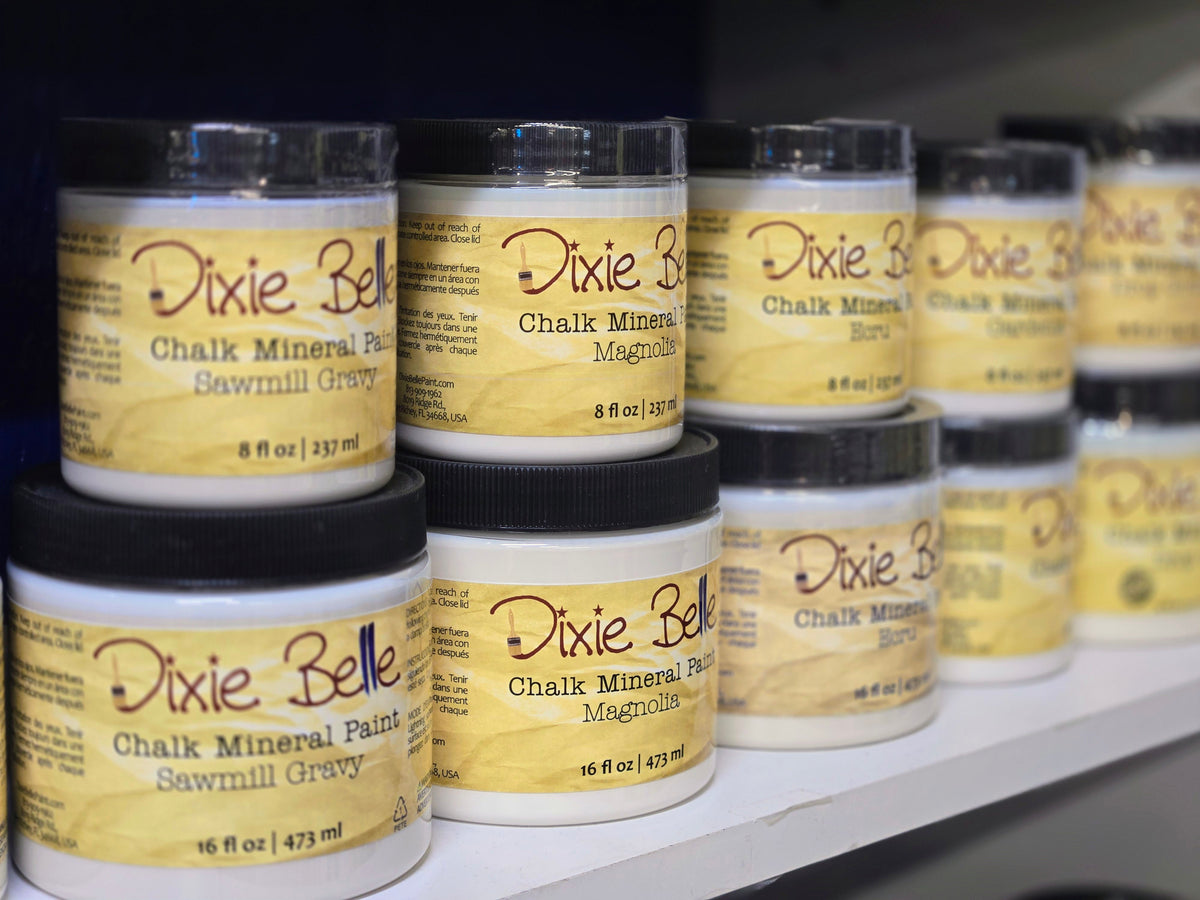 Several Dixie Belle Chalk Mineral Paint jars stacked on a shelf, with the front labels clearly showing 'Dixie Belle' and 'Chalk Mineral Paint' in a warm yellow-and-brown design.
