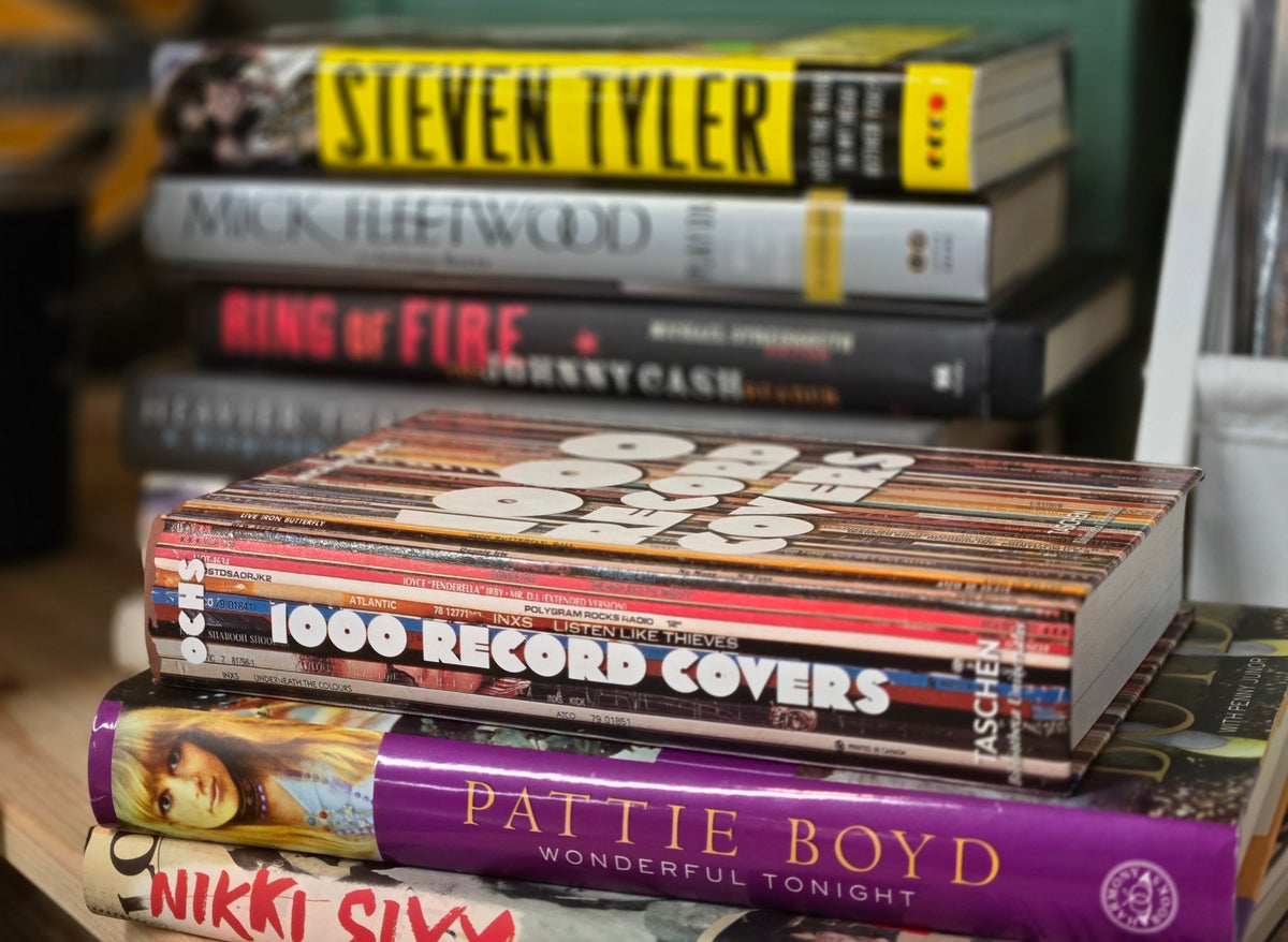 A close-up stack of books on a shelf, with the front-most title visible as '1000 Record Covers' and other visible authors including Steven Tyler, Mick Fleetwood, Ring of Fire, Johnny Cash, Pattie Boyd, and Nikki Sixx.