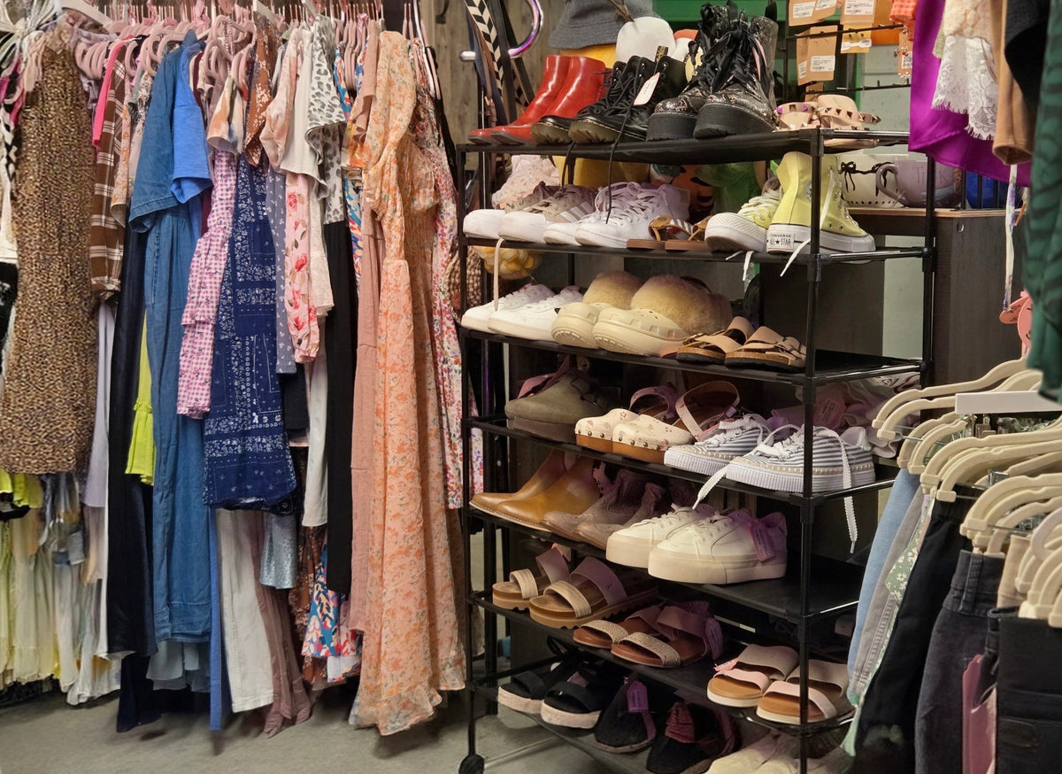 A retail clothing and shoe display with racks of colorful dresses and tops on the left, a black shelving unit filled with sneakers, sandals, boots, and shoes in the center-right, and more garments hanging on the right.