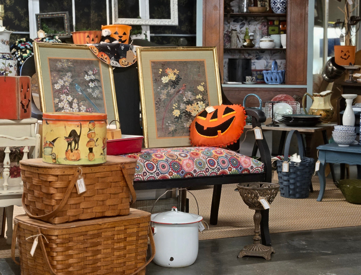 A styled indoor display featuring two large framed floral art prints with gold frames, a patterned upholstered bench with a jack-o'-lantern pillow, wicker baskets, vintage tins, and other home decor items in a shop-like setting.