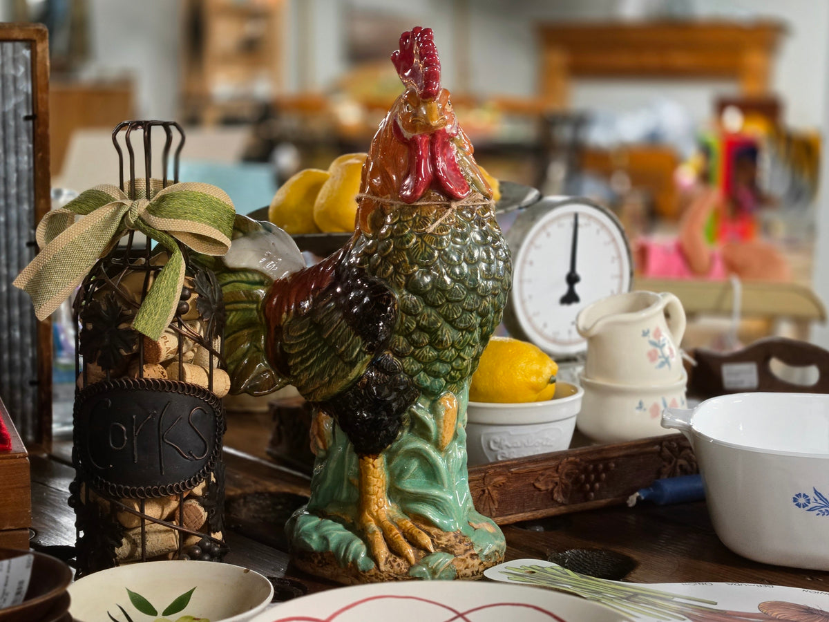 A decorative ceramic rooster figurine in green, black, red, and yellow glaze, displayed on a rustic table alongside a wire bottle rack with a ribbon bow, lemons, a small cream pitcher, and bowls in a softly blurred indoor setting.