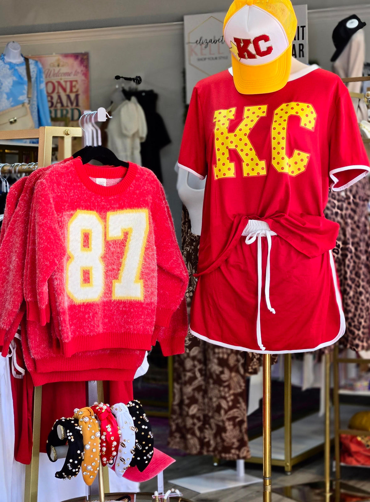 A retail display featuring a red crewneck sweater with a large yellow and white '87' on the front, and a matching red lounge set on a mannequin with a yellow and white 'KC' baseball cap. The lounge set includes a short-sleeve top with 'KC' lettering and a matching tie-waist shorts set.