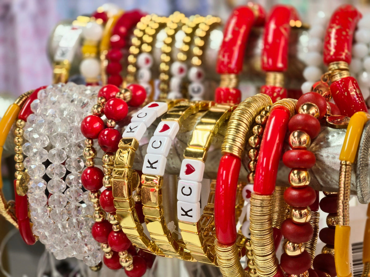 A close-up display of assorted bangles and bracelets, featuring gold-plated chain and link styles, red and white beads, clear faceted crystal strands, and letter tiles spelling KC and AK on some bracelets.