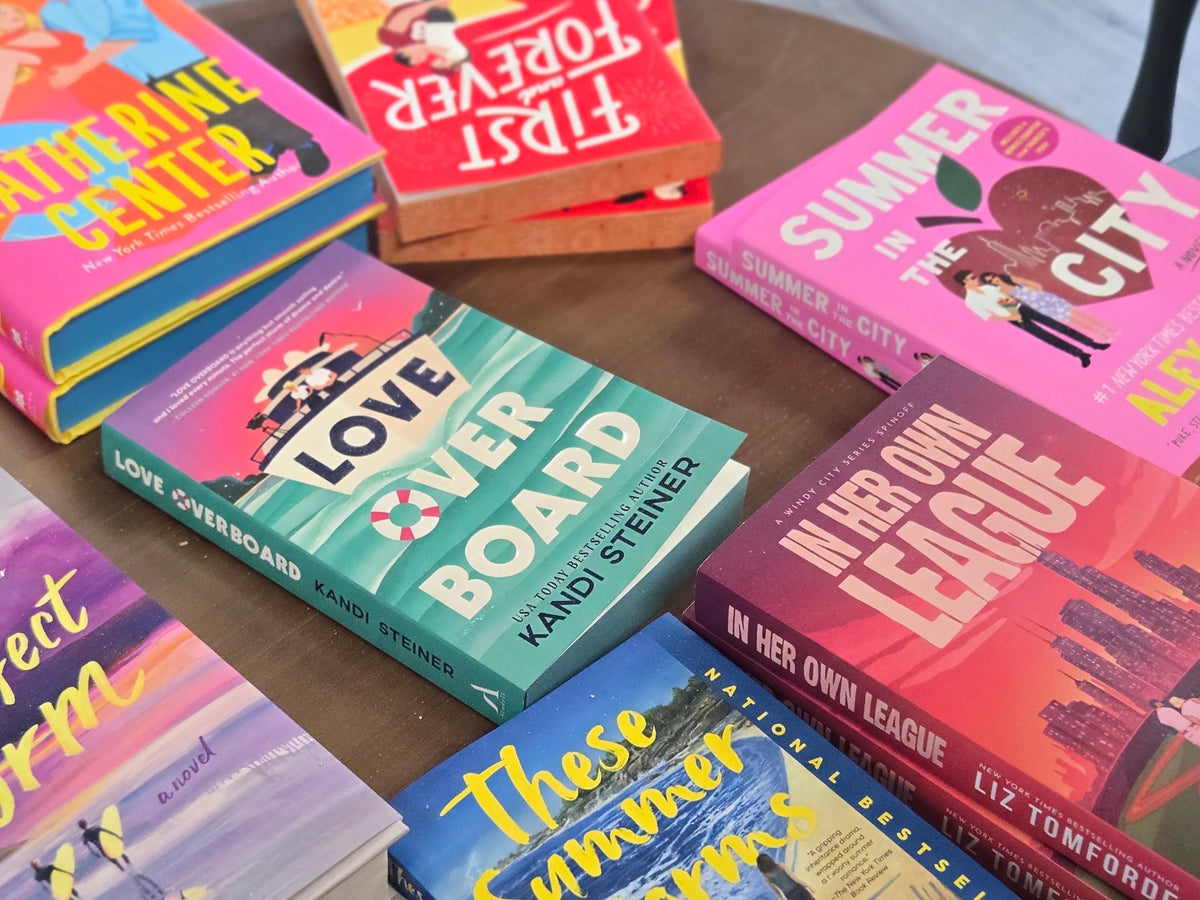 A group of colorful romance novel books arranged on a wooden table, including titles such as Love Board, Summer in the City, and These Summer Nights.