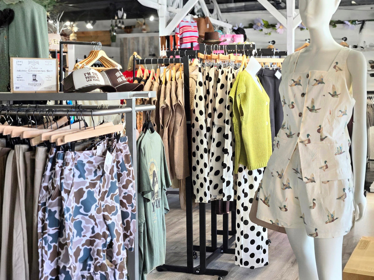 A boutique clothing display featuring a white mannequin wearing a sleeveless dress with a small bird print, alongside racks of patterned garments including polka dot and camouflage styles.