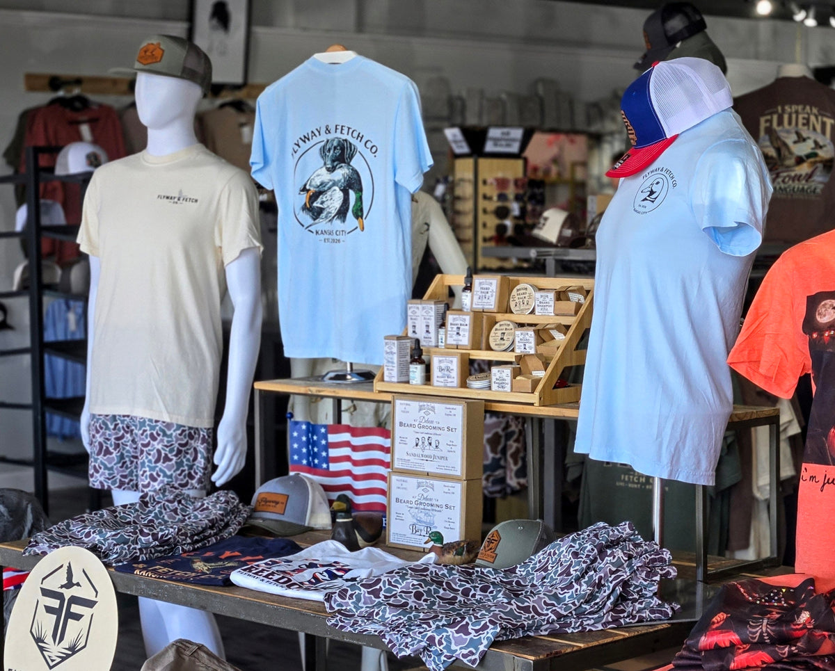 A retail store display featuring mannequins wearing graphic t-shirts and patterned shorts, with a central wooden table holding small boxes, a U.S.