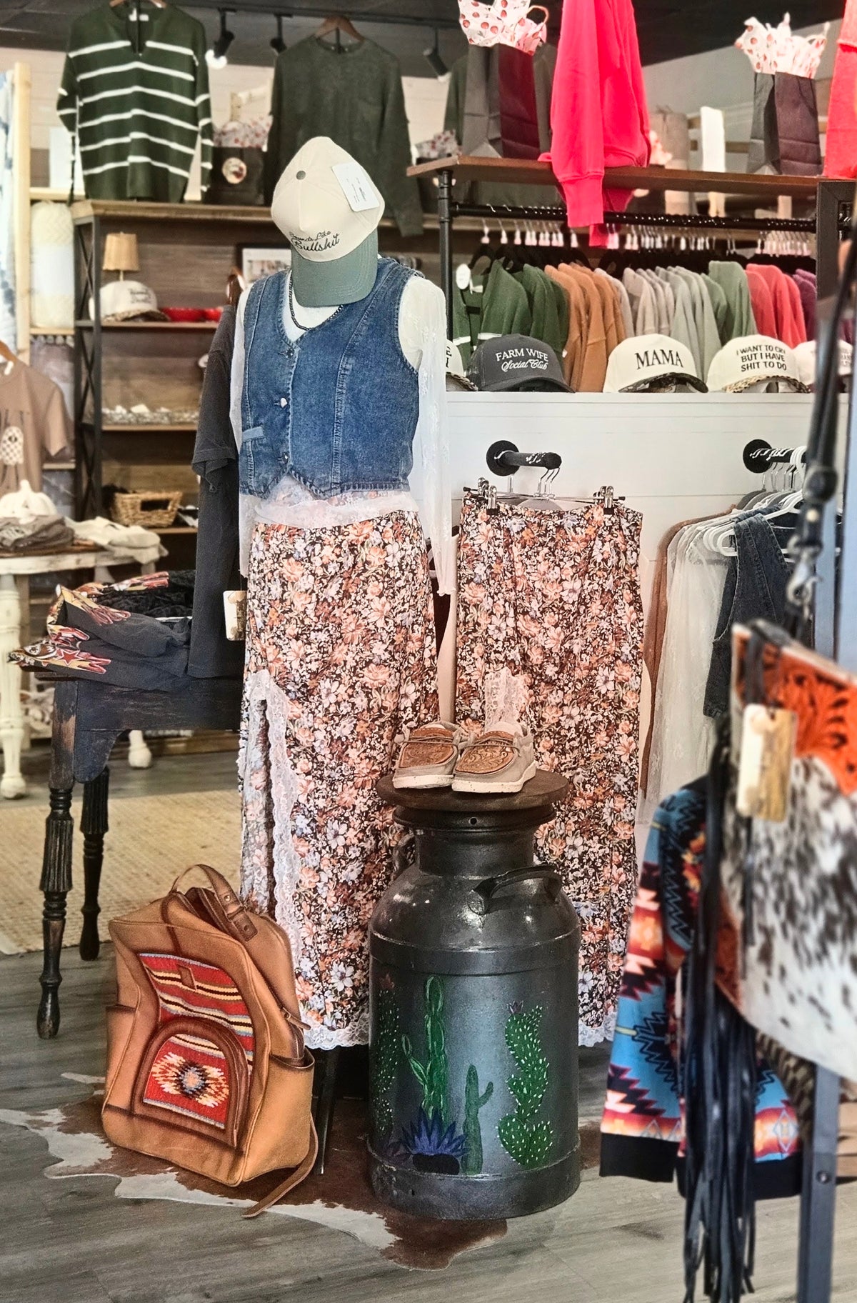A styled clothing display inside a boutique featuring a mannequin dressed in a denim vest, white top, and floral wide-leg pants, with matching floral pants hanging on a rack and a brown patterned backpack beside a decorative black metal canister.