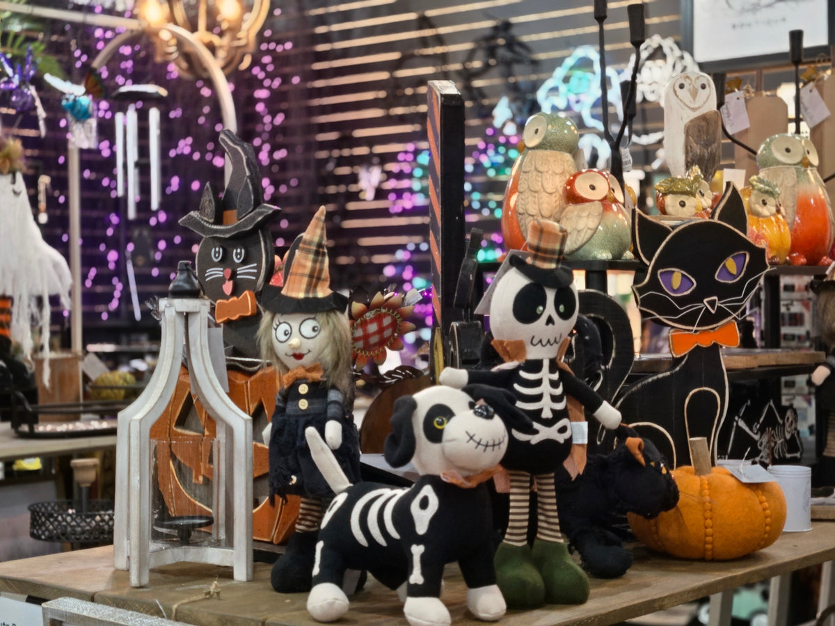 A display of various Halloween decorations on a wooden table, including plush toys, cardboard cutouts, a skeleton dog, a witch scarecrow, and a black cat with a pumpkin, set against a busy shop background with lights.