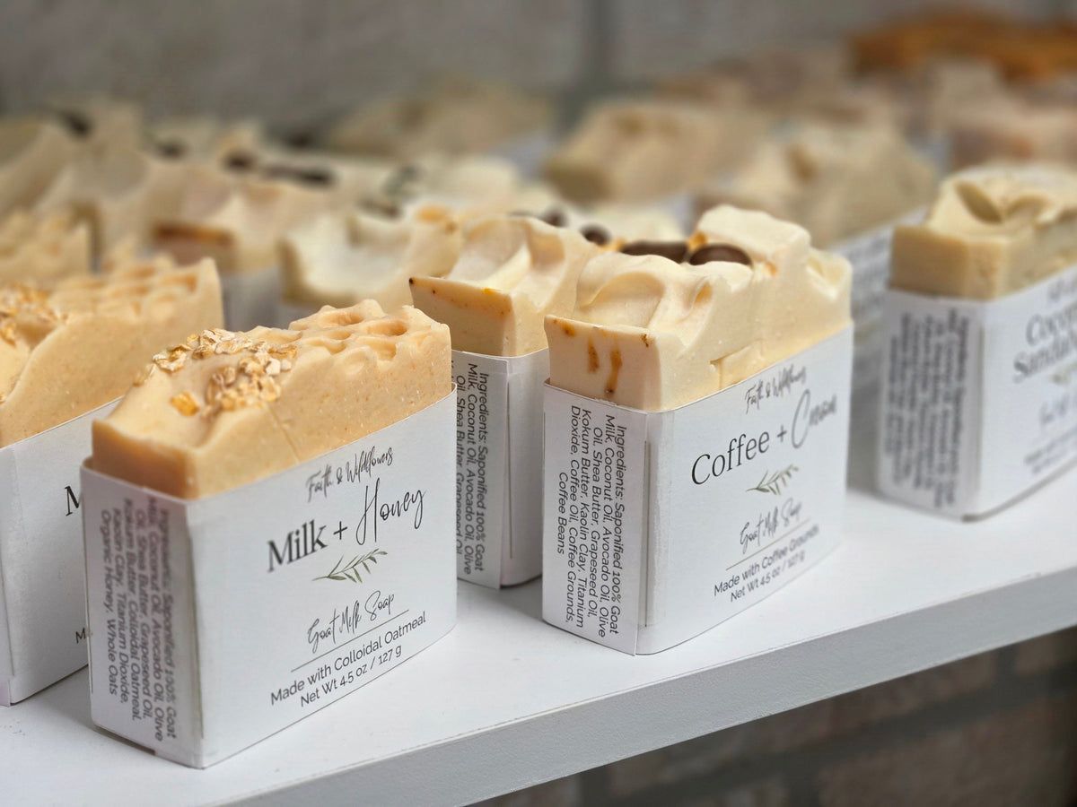 Several handmade soap bars in white paper sleeves are arranged on a white shelf. The soaps have light beige to tan tones with some darker brown swirls and toppings, and the labels read Milk + Honey, Coffee + Cinnamon, and others.